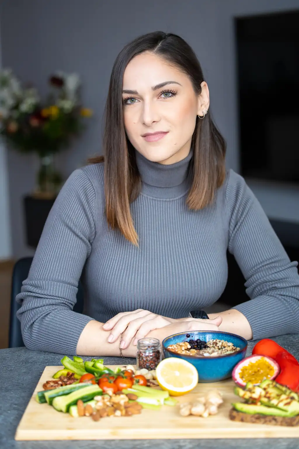 Andreea Petre in grauem Rollkragenpullover sitzt aufrecht am Tisch, die Hände locker übereinandergelegt, und blickt mit leichtem Lächeln in die Kamera. Vor ihr ein Holzbrett mit Gurke, Staudensellerie, Kirschtomaten, Nüssen und Zitrone sowie eine blaue Schale mit Körner- und Saatenmischung; im unscharfen Hintergrund ein Blumenstrauß.