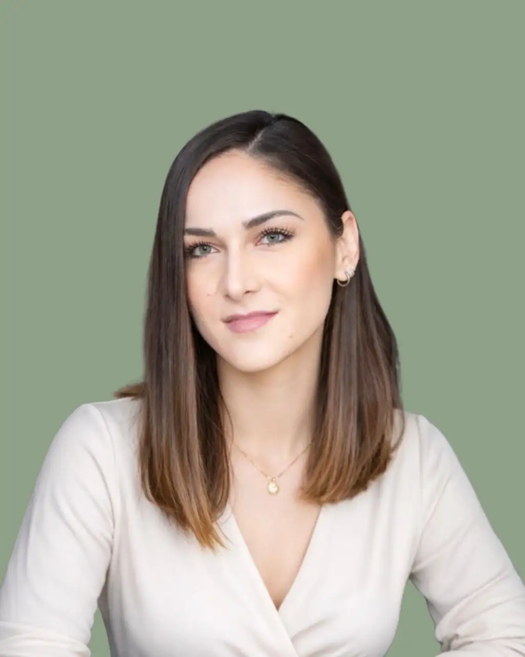 Andreea Petre, Trainerin für metabolische Migräne-Therapie, in cremefarbener Wickelbluse mit feiner Halskette blickt mit ruhigem, freundlichem Ausdruck direkt in die Kamera. Ihr langes dunkelbraunes Haar fällt offen über die Schultern; im Hintergrund eine schlichte salbeigrüne Fläche.