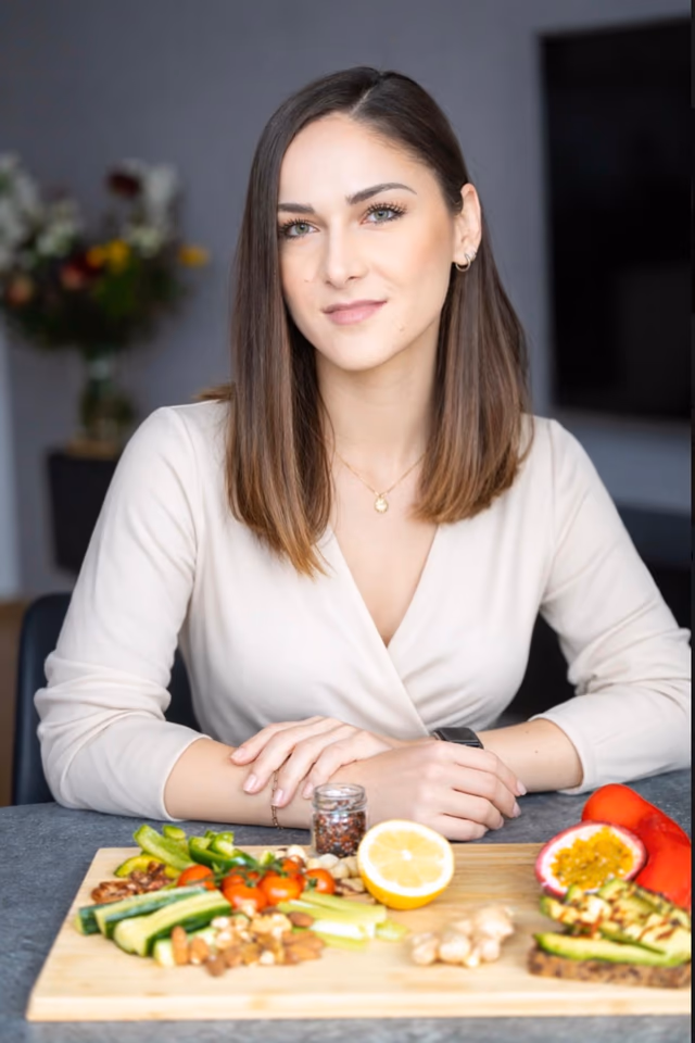 Andreea Petre, Trainerin für metabolische Migräne-Therapie, sitzt aufrecht am Tisch und blickt ruhig in die Kamera.